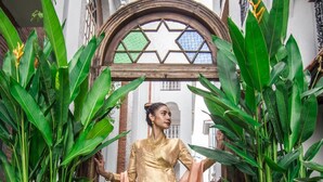 Una persona vestida con traje tradicional se encuentra en un patio con un edificio blanco, plantas verdes grandes y un arco decorativo.