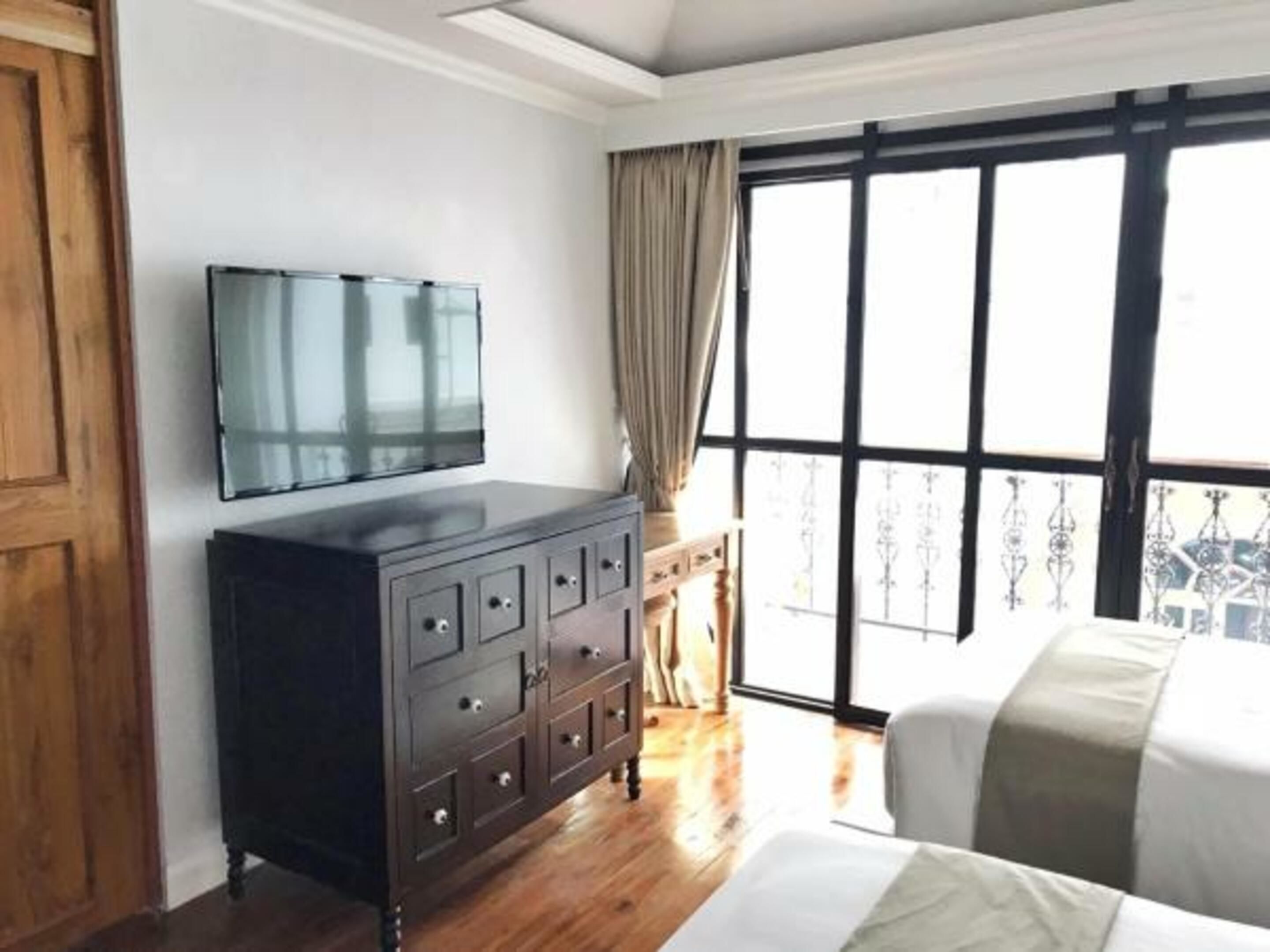 Una habitación de hotel con un ventanal grande, un televisor de pantalla plana sobre una cómoda de madera oscura y una cama con edredón blanco y gris a rayas.
