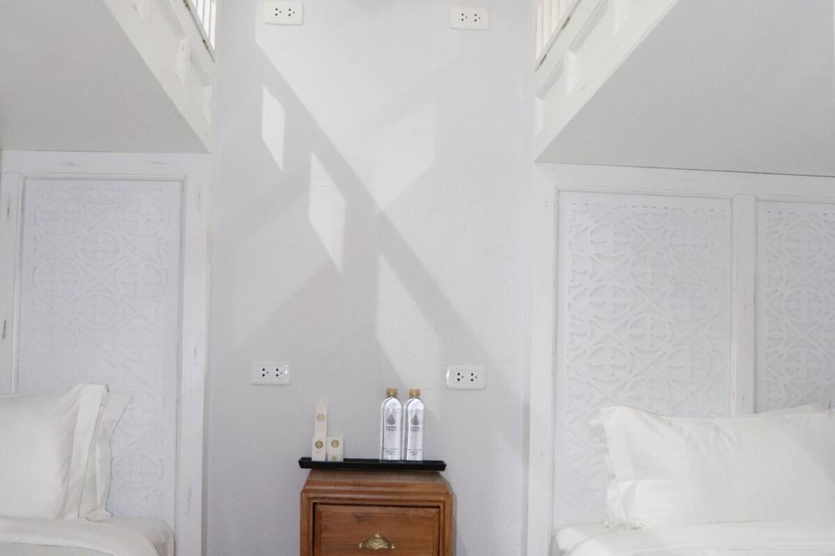A narrow room with two beds, a wooden nightstand, and a small shelf above the nightstand.