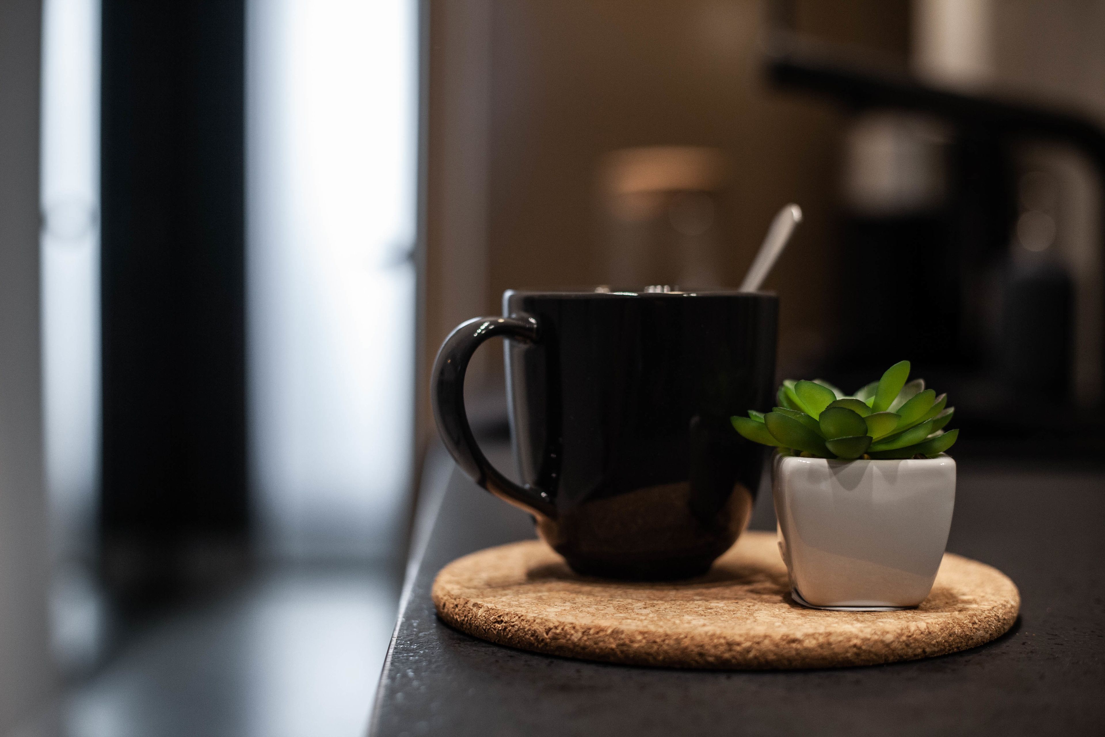 A black mug with a spoon, a small white pot with a green plant, and a cork coaster on a dark countertop.