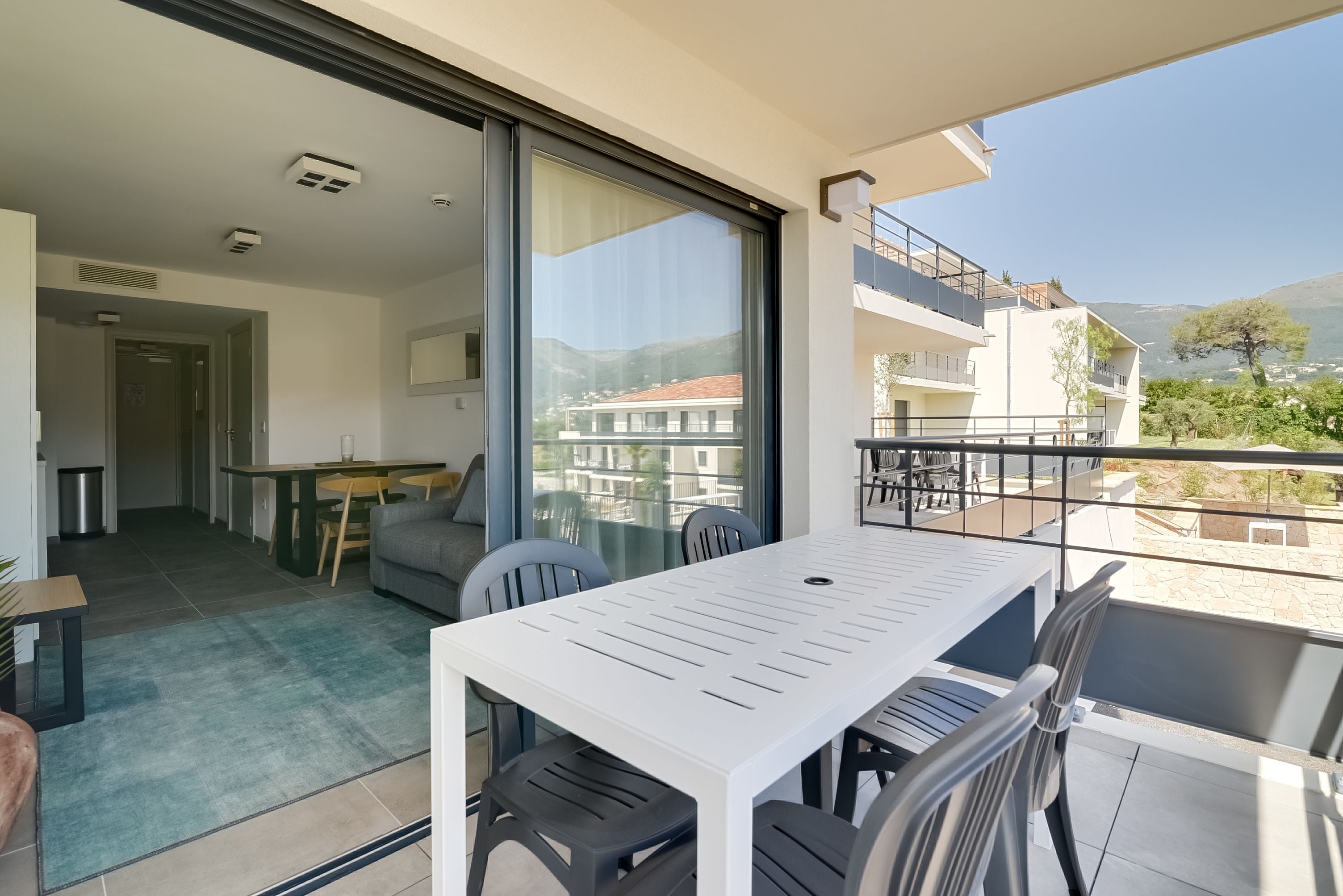 Een balkon met een tafel en stoelen, een glazen schuifdeur die leidt naar een woonruimte en uitzicht op gebouwen en heuvels.