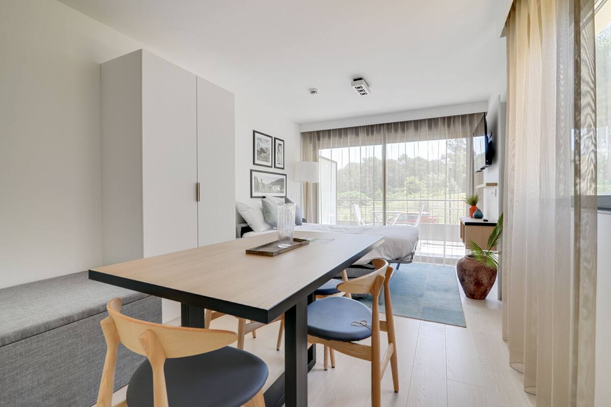 A modern, well-lit room with a dining table, chairs, a sofa, and a large window offering a view of greenery.