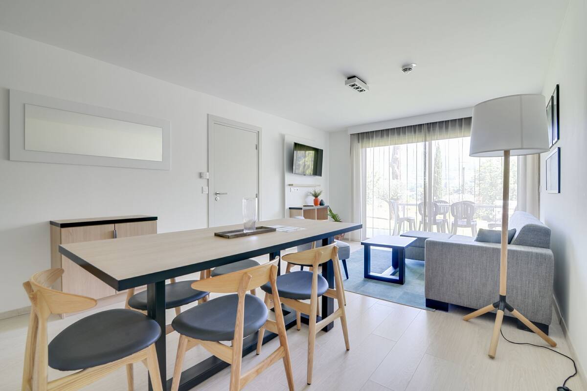 A modern kitchen with a dining area, featuring a long table, wooden chairs, and a grey sofa.