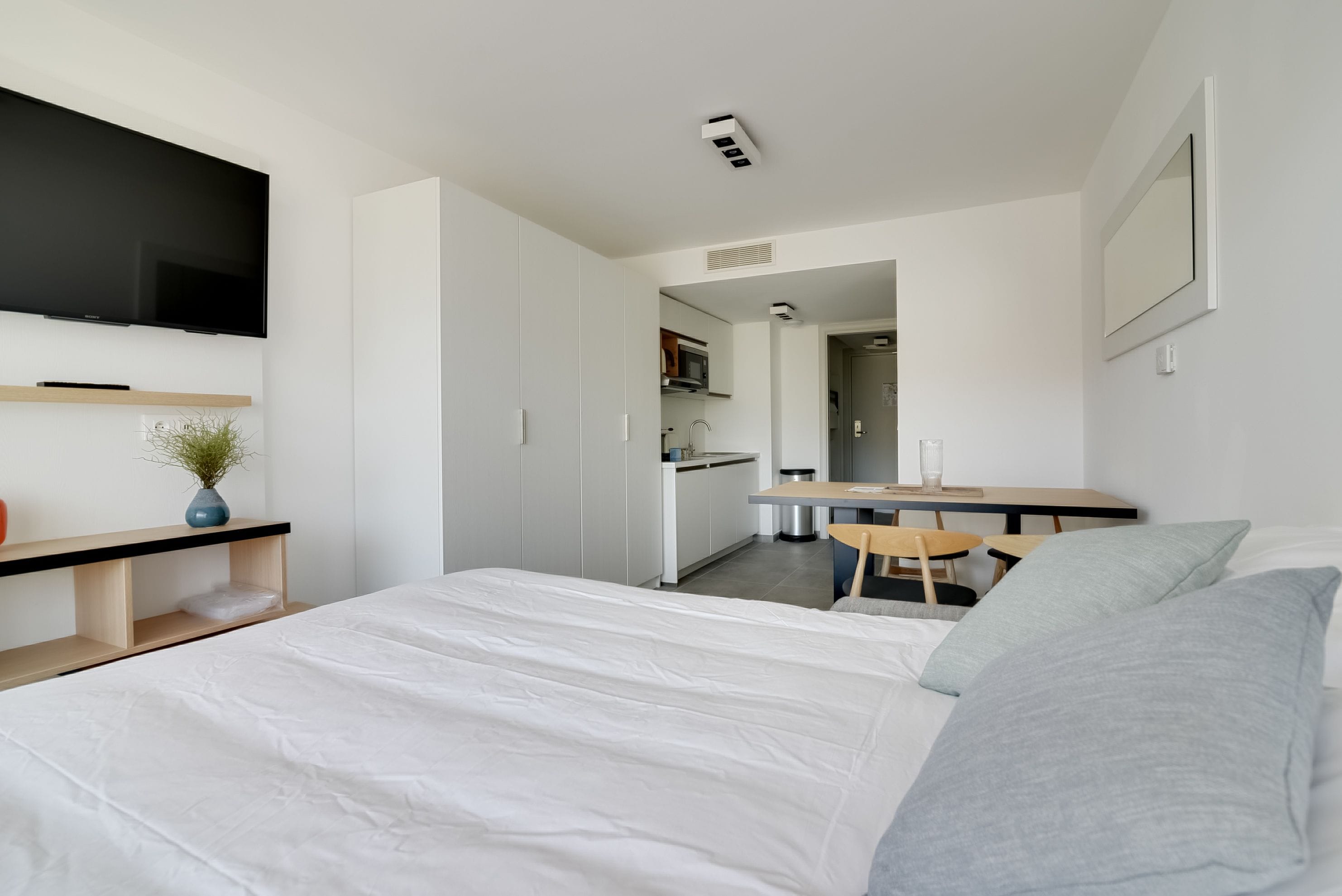 A modern hotel room with a large bed, a desk, a kitchenette, and a TV mounted on the wall.