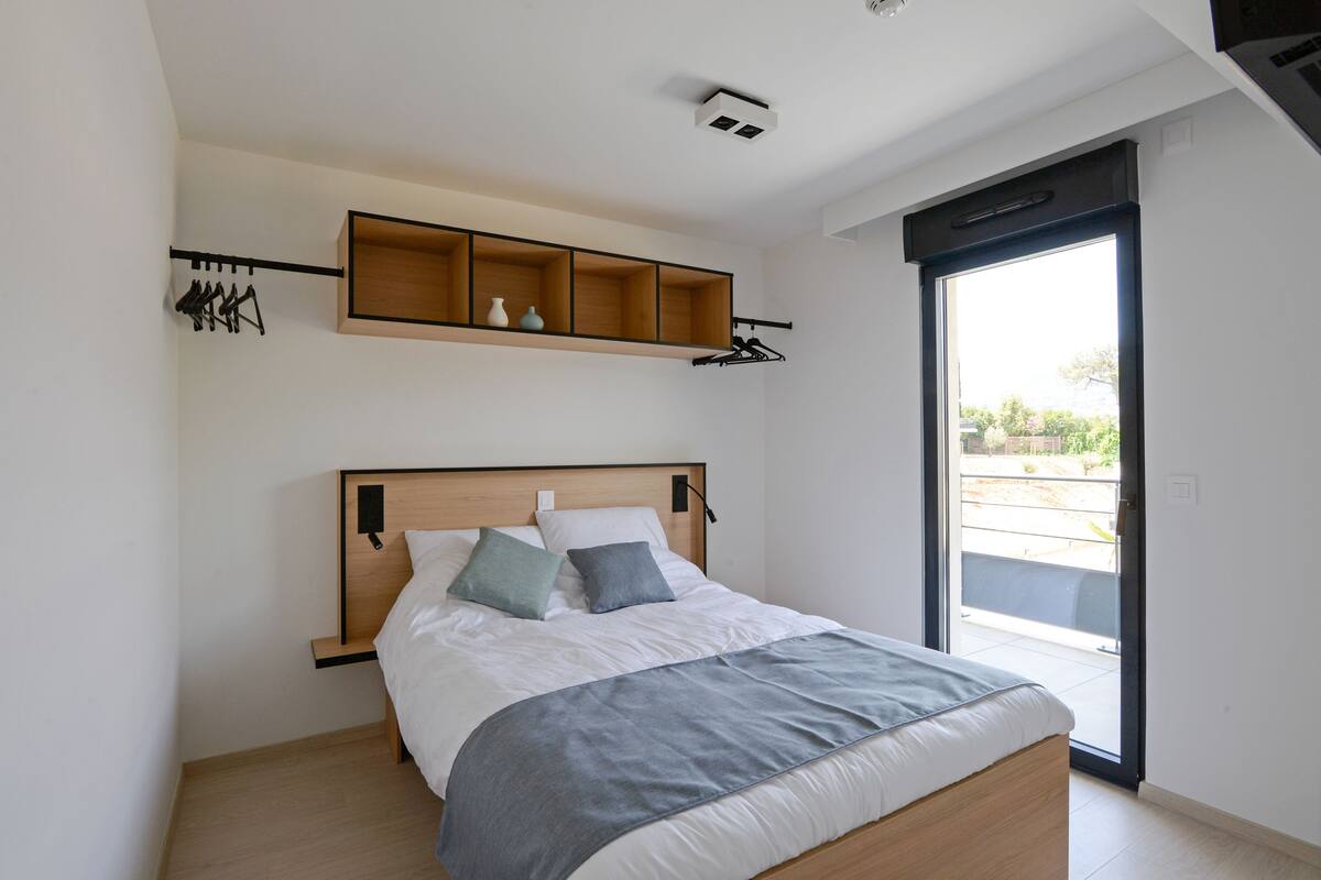 A bedroom with a bed, a built-in shelf above the bed, a coat rack, and a sliding glass door to the outside.