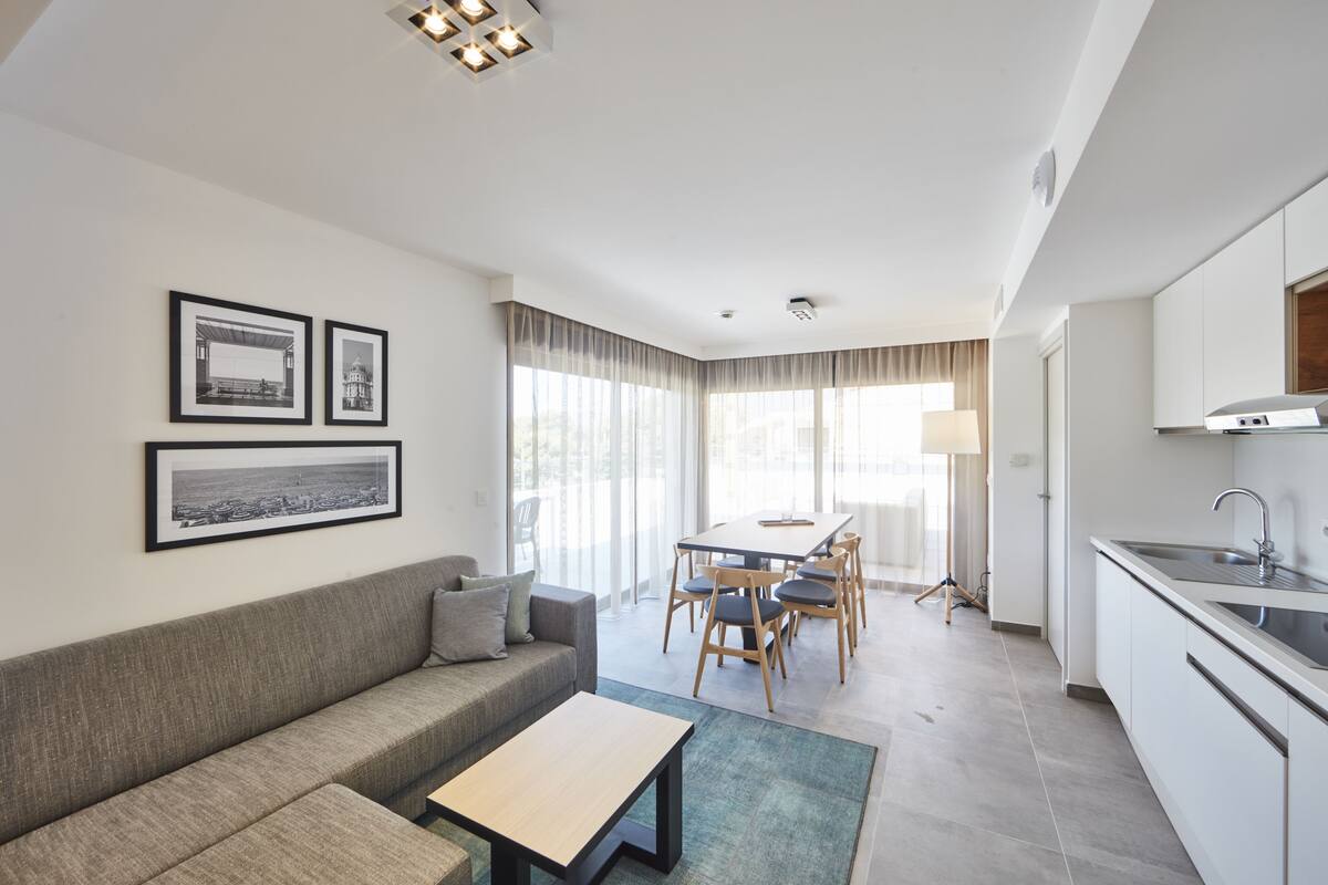 A modern living room with a sofa, coffee table, and dining area with chairs. There is a kitchen with a sink and cabinets.