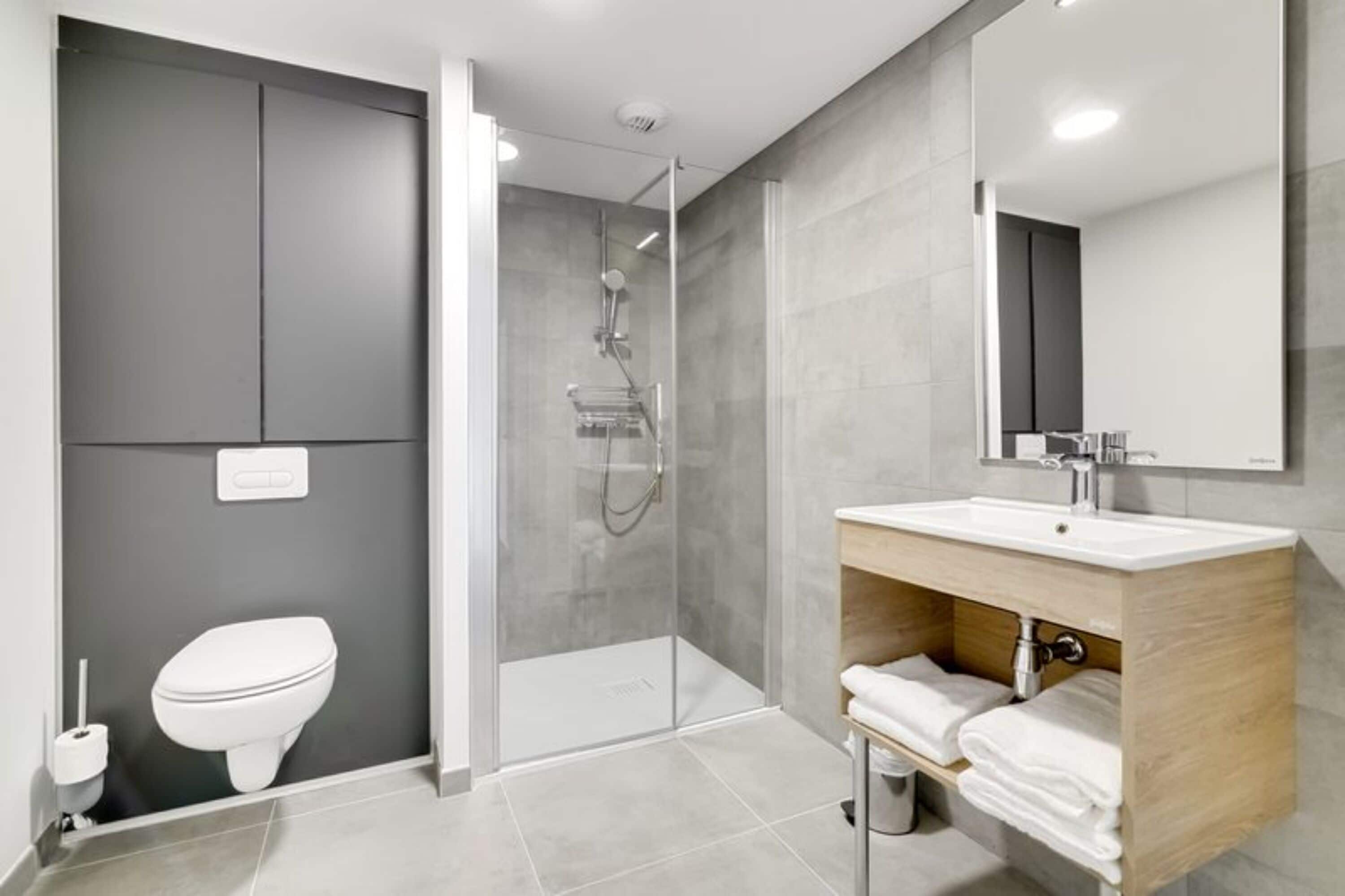 A modern bathroom with a glass shower enclosure, a white toilet, and a wooden vanity with a sink and towel rack.