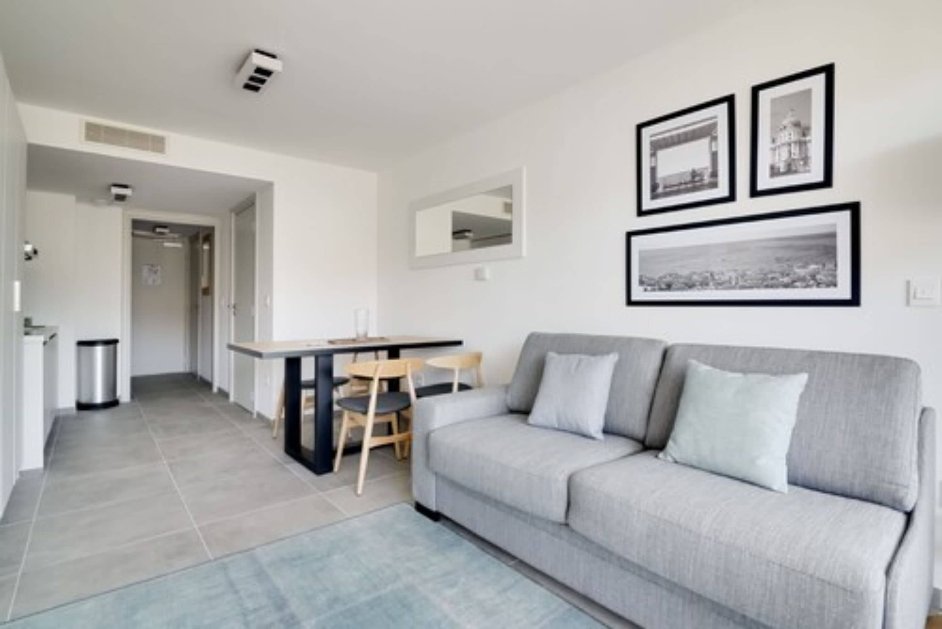 A modern living room with a grey sofa, a dining table with chairs, and framed pictures on the wall.