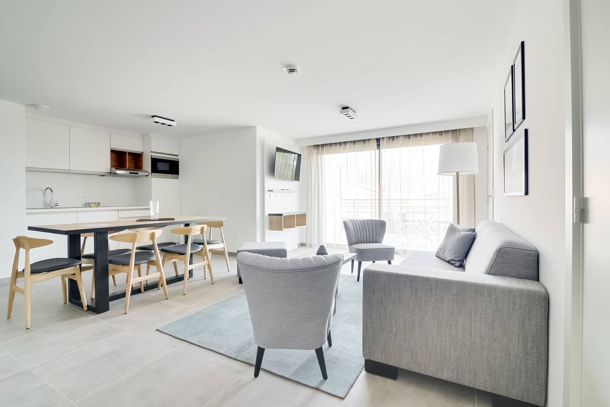 A modern living room with a grey sofa, a dining area with wooden chairs, and a kitchen with white cabinets.