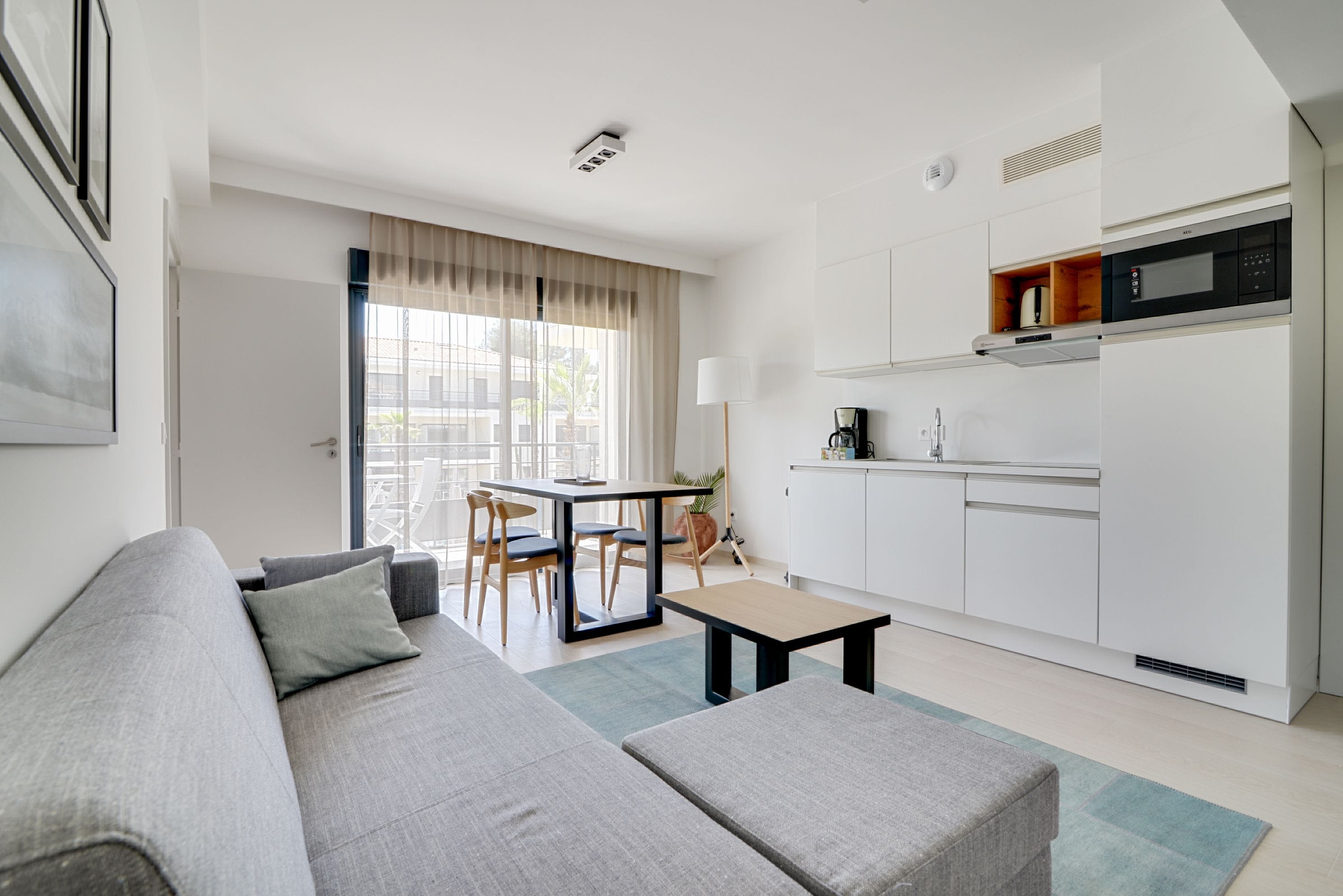 Un salon moderne avec un canapé gris, une petite table basse et un coin repas comprenant une table et des chaises. On aperçoit, au fond, une cuisine avec des meubles blancs et un four à micro-ondes.