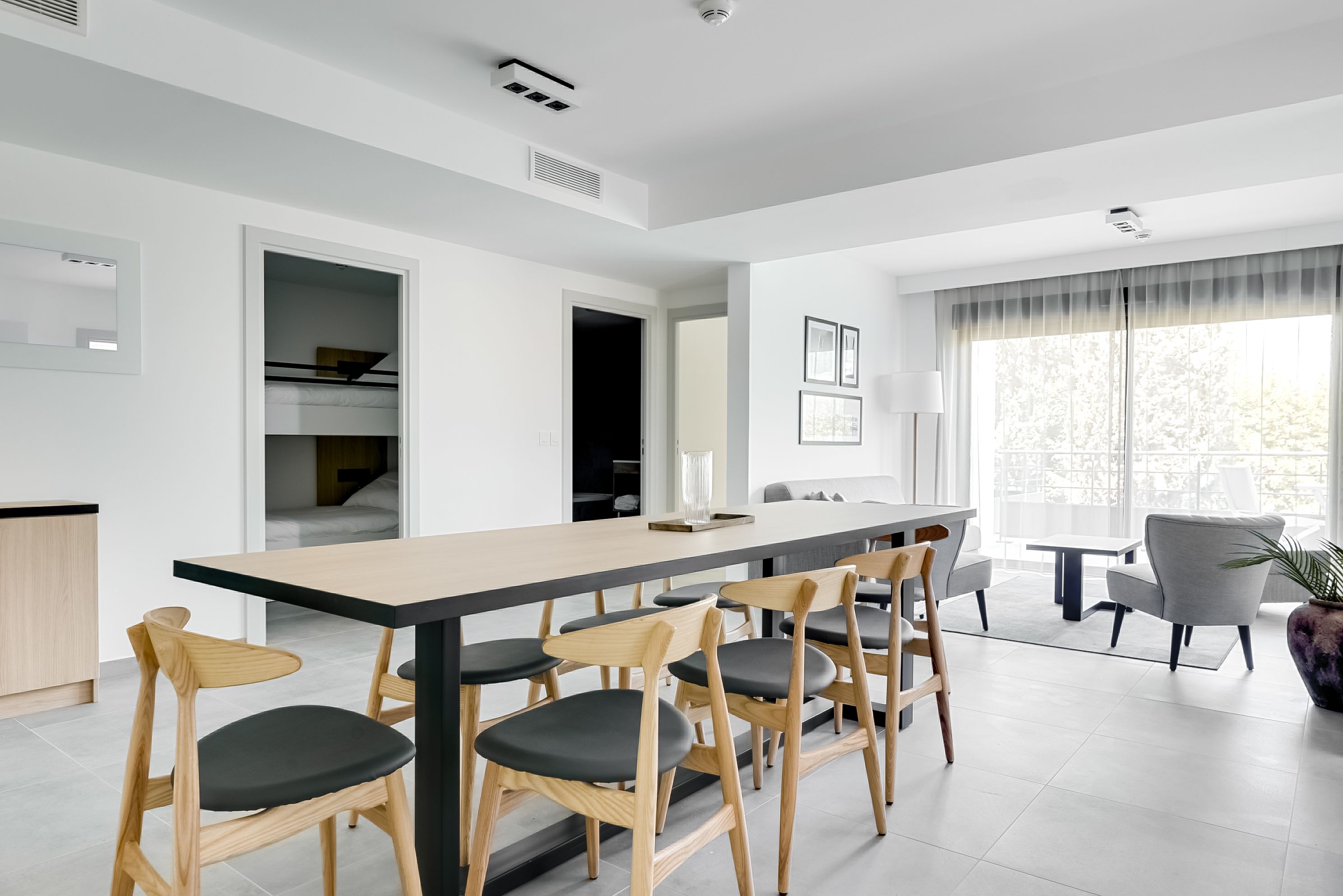 A modern dining area with a long table and six wooden chairs with black cushions. There is a small table with a vase on the table, and a grey armchair in the background.