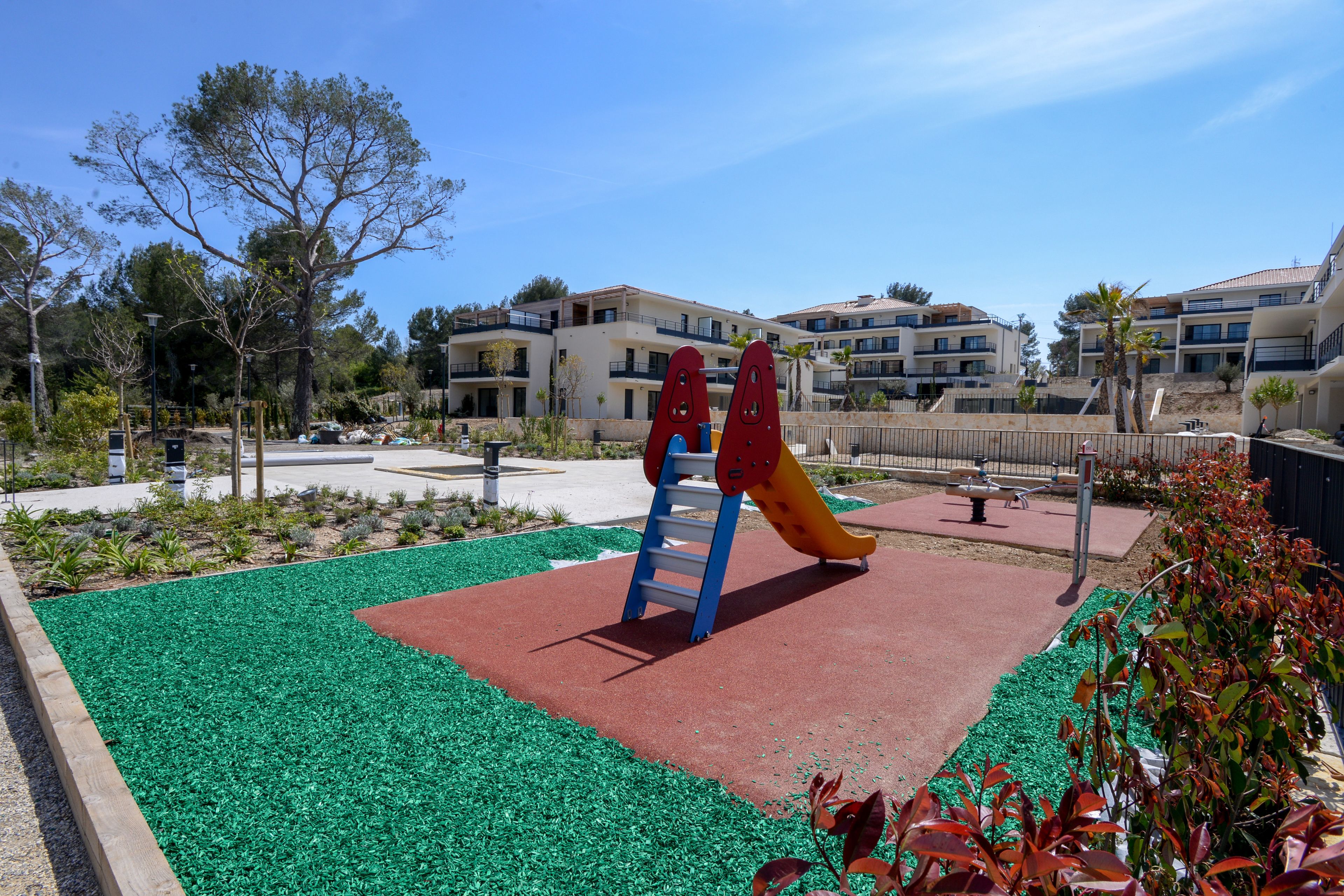 Un parque infantil con tobogán y estructura de escalada, rodeado de césped artificial verde y suelo de goma roja, con edificios residenciales al fondo.