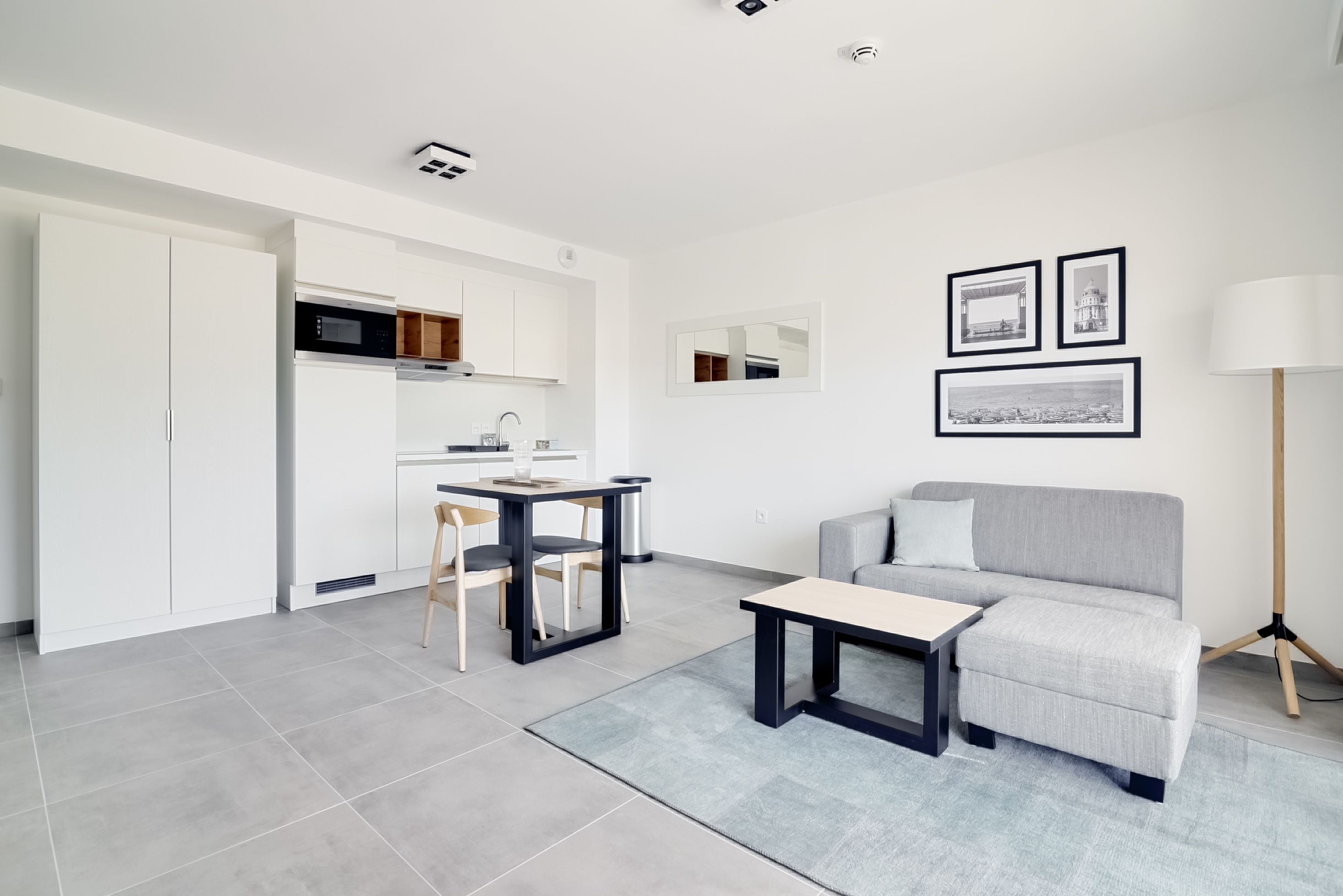 Een moderne woonkamer met een grijze bank, een klein tafeltje en een eethoek met een tafel en stoelen. Er hangen ingelijste foto's aan de muur en staat een lamp naast de bank.