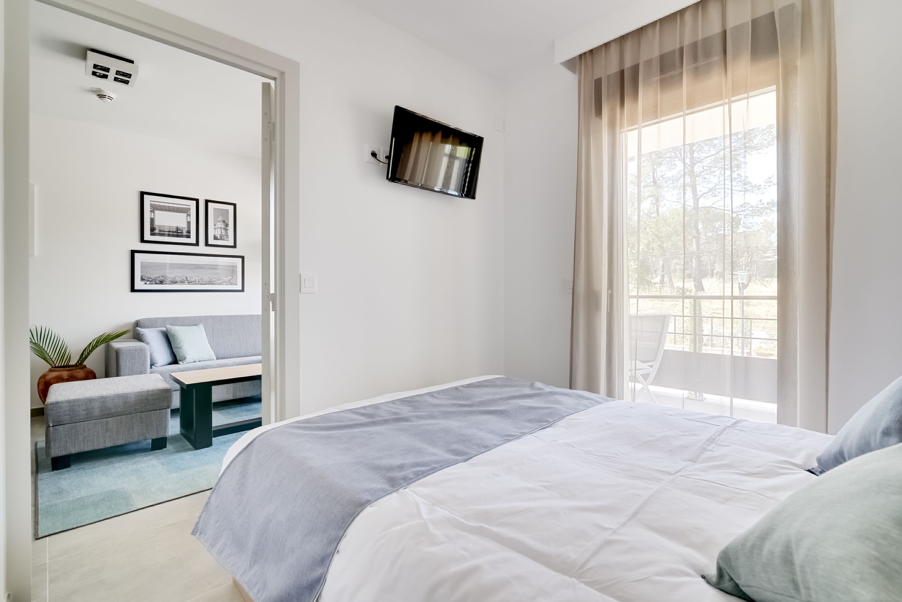 A modern bedroom with a bed, a sofa, and a small table. There are framed pictures on the wall and a window with curtains.