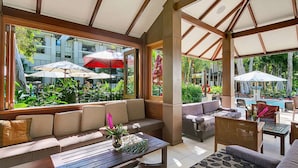 A sunlit indoor patio with a sofa, dining table, and umbrellas, overlooking a pool and lush greenery.