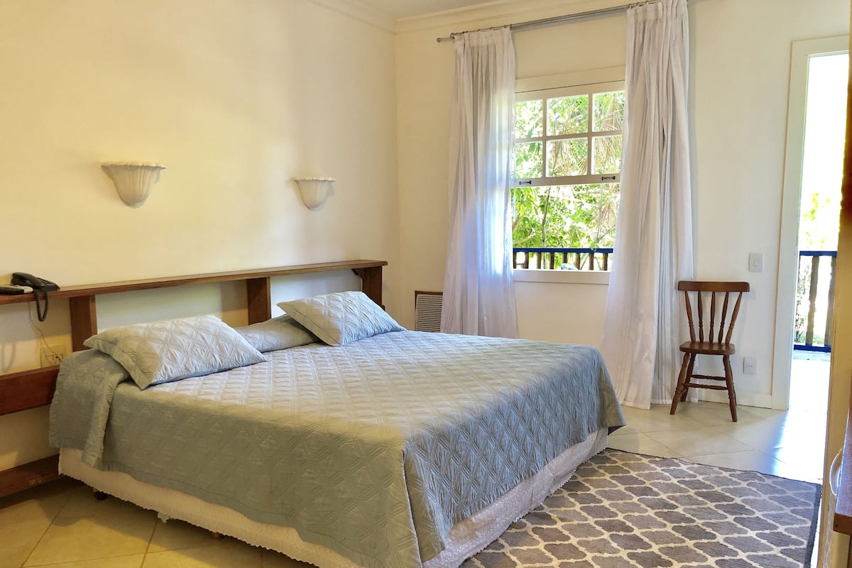 Une chambre à coucher avec un lit, des lampes de chevet, une fenêtre avec des rideaux, une chaise et une vue sur la verdure à l’extérieur.