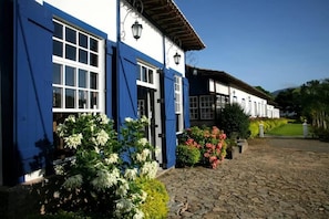 Un edificio con fachada blanca y contraventanas azules, un camino de adoquines y un jardín cuidado con diversas plantas y flores.