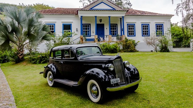 Un coche antiguo negro estacionado frente a una casa blanca con detalles azules y columnas.