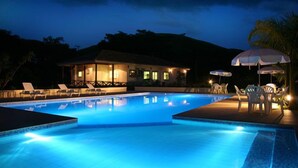Una piscina nocturna con sillones de descanso y sombrillas, rodeada por un edificio iluminado.