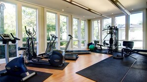 Un gimnasio amplio con diversos equipos de ejercicio, ventanas grandes y vistas al exterior con vegetación.