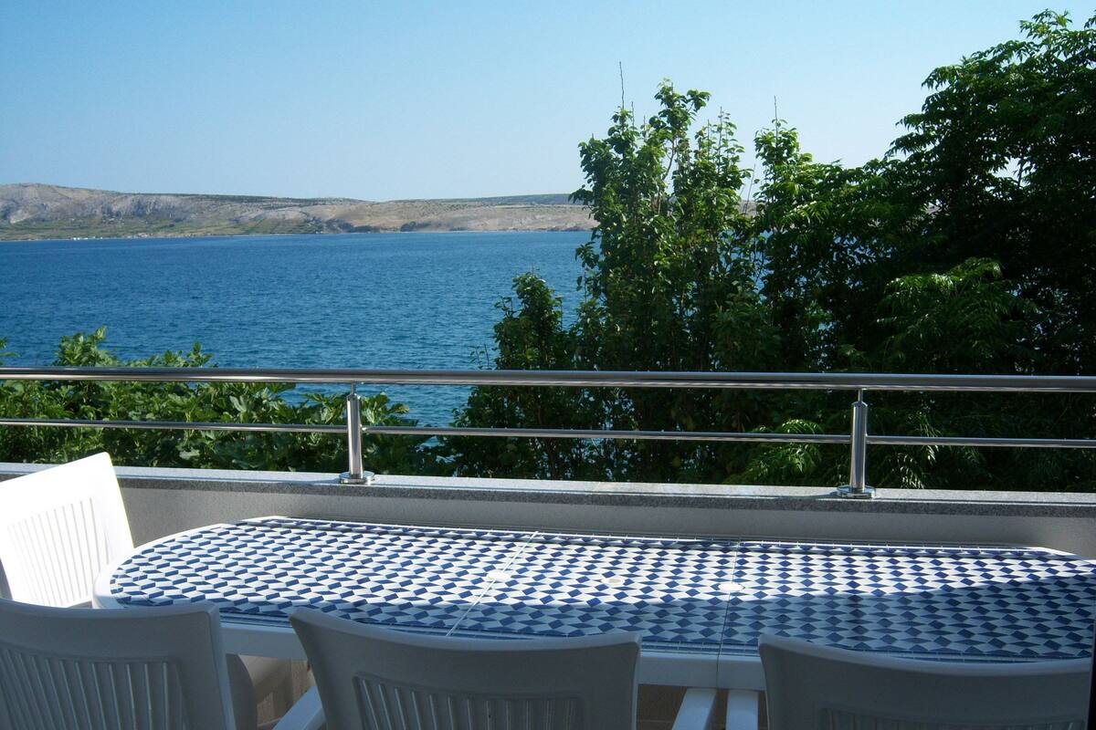 Een tafel met een blauw-wit geruit tafelkleed, omringd door witte plastic stoelen, met uitzicht op een waterpartij en heuvels in de verte.