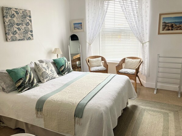Un dormitorio ordenado con una cama, dos sillones, un espejo y obras de arte en las paredes.