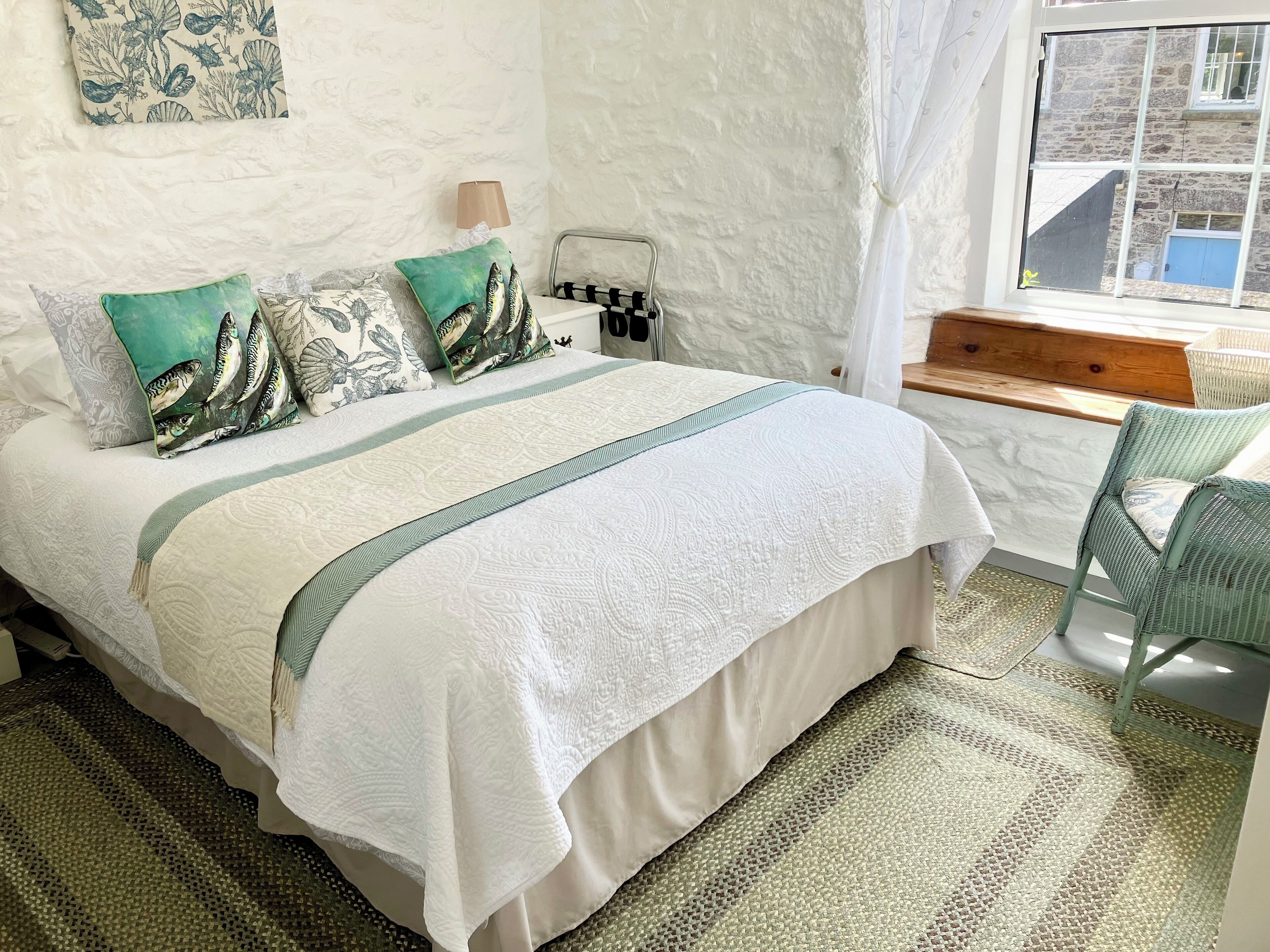A bed with a white and green bedspread, two pillows with fish designs, a bedside table with a lamp, and a view of a stone building through a window.