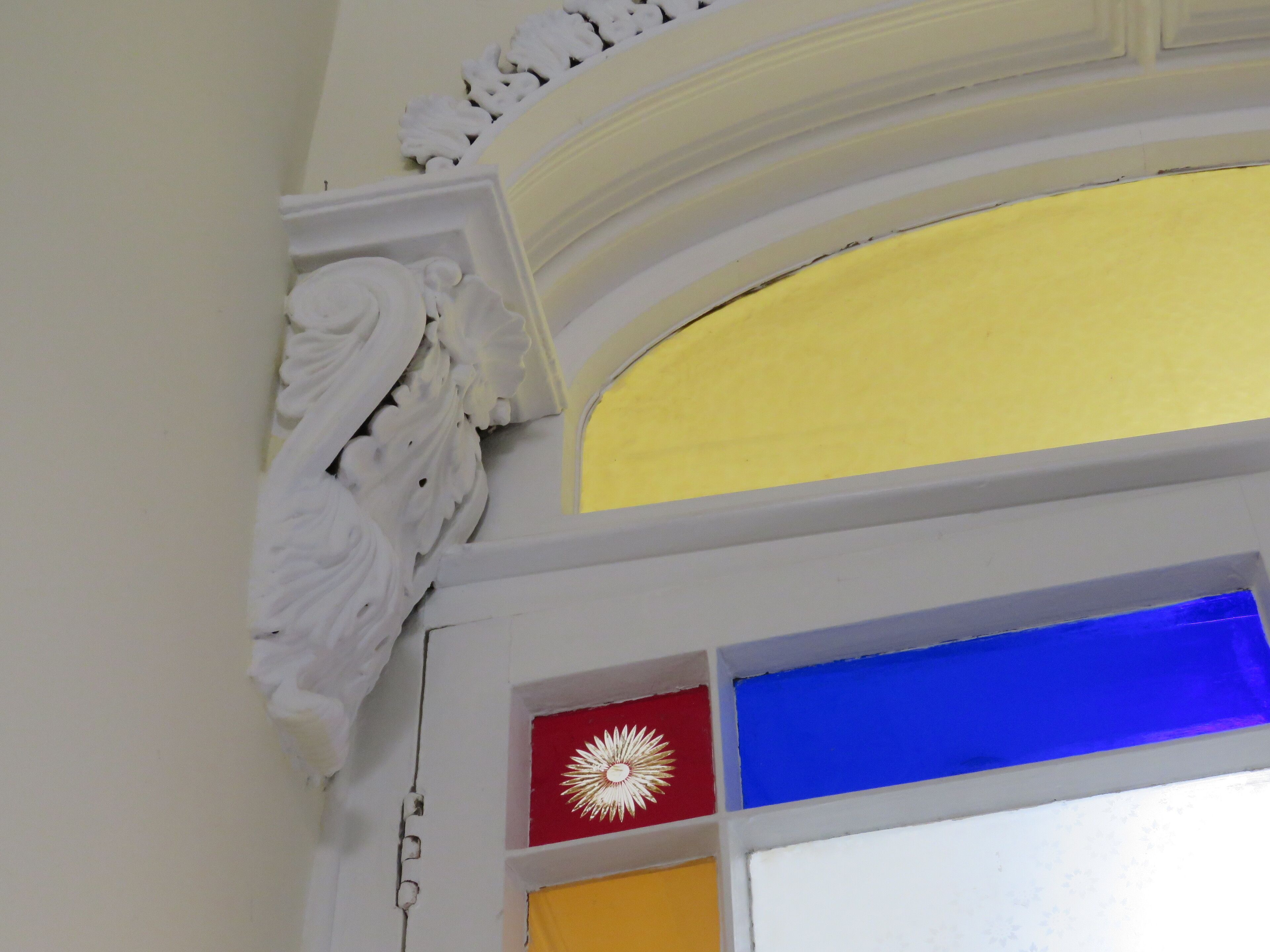 Parte del interior de un edificio con molduras blancas decorativas, un arco pintado de amarillo y azul, y un panel rojo con un diseño similar a un sol blanco.