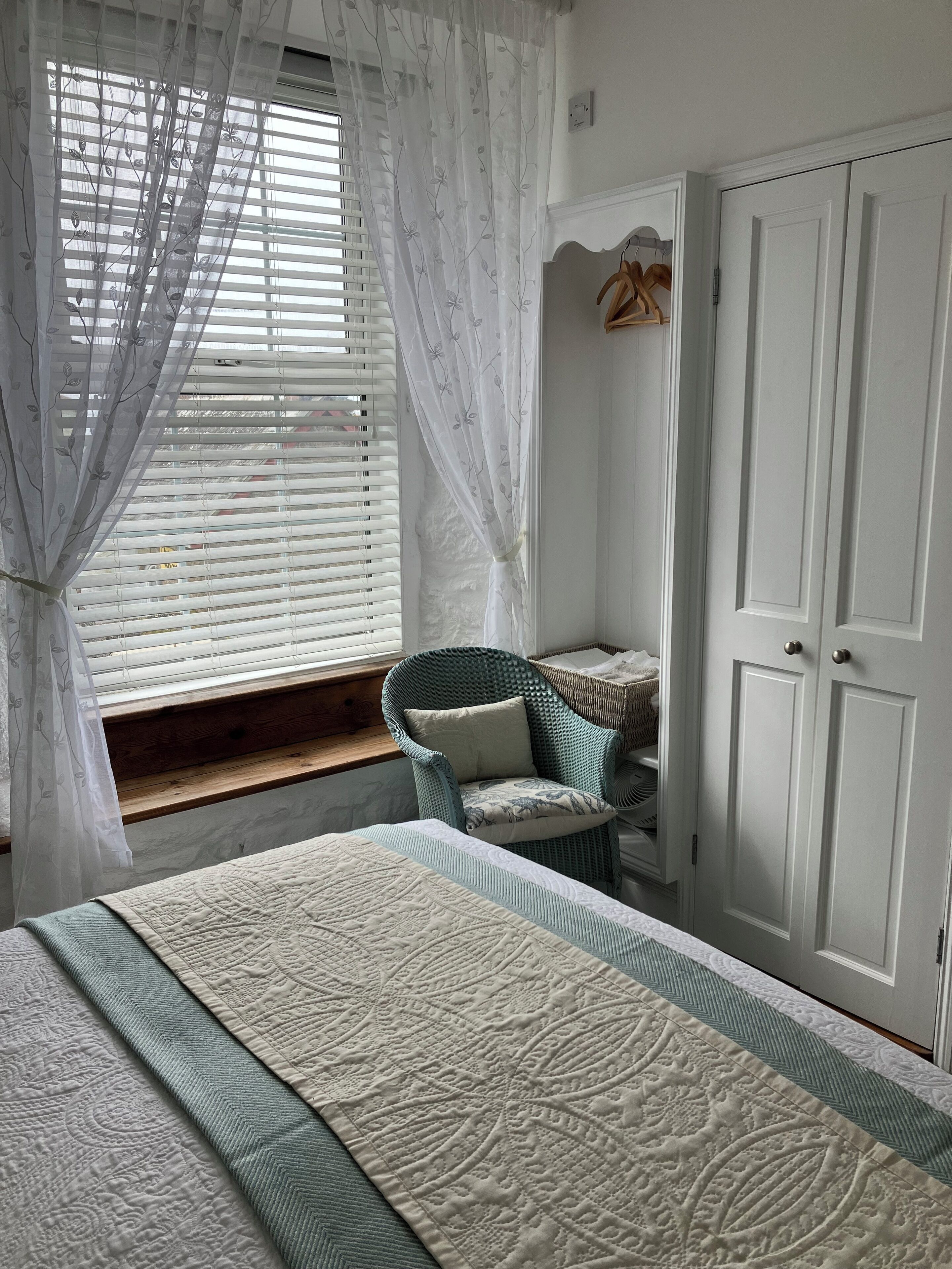 A bedroom with a bed, a wicker chair, a wardrobe, and a window with sheer curtains.