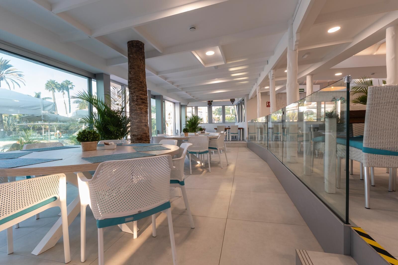 Un comedor moderno con mobiliario de mimbre blanco, una columna central y grandes ventanales con vistas a palmeras.
