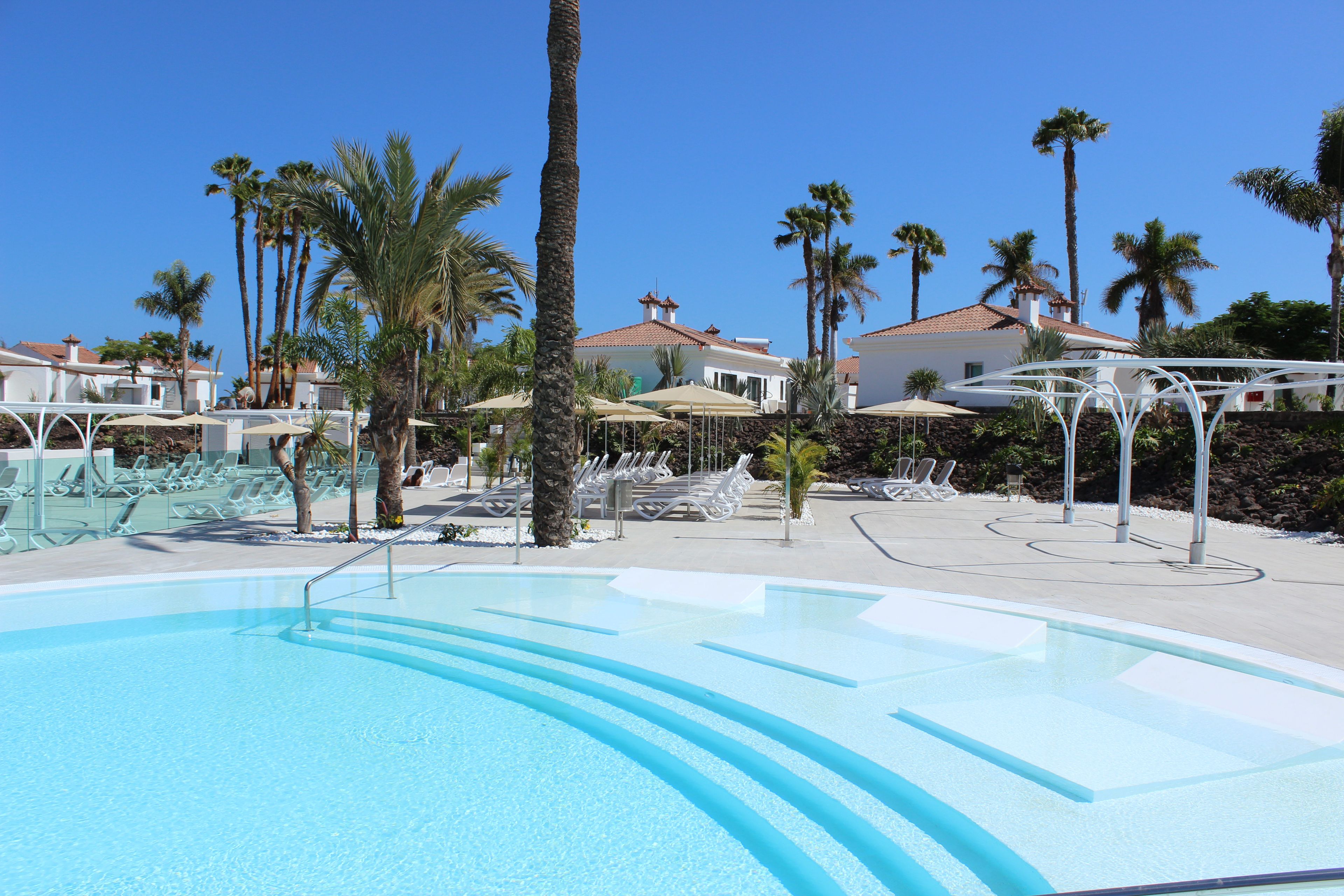 Una piscina con sillas de descanso y sombrillas, rodeada de palmeras y edificios.