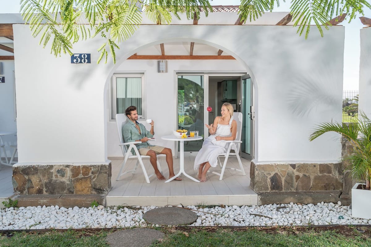 Un couple est assis à une table sur une terrasse, devant un mur de pierre et un panneau indiquant le numéro 38.
