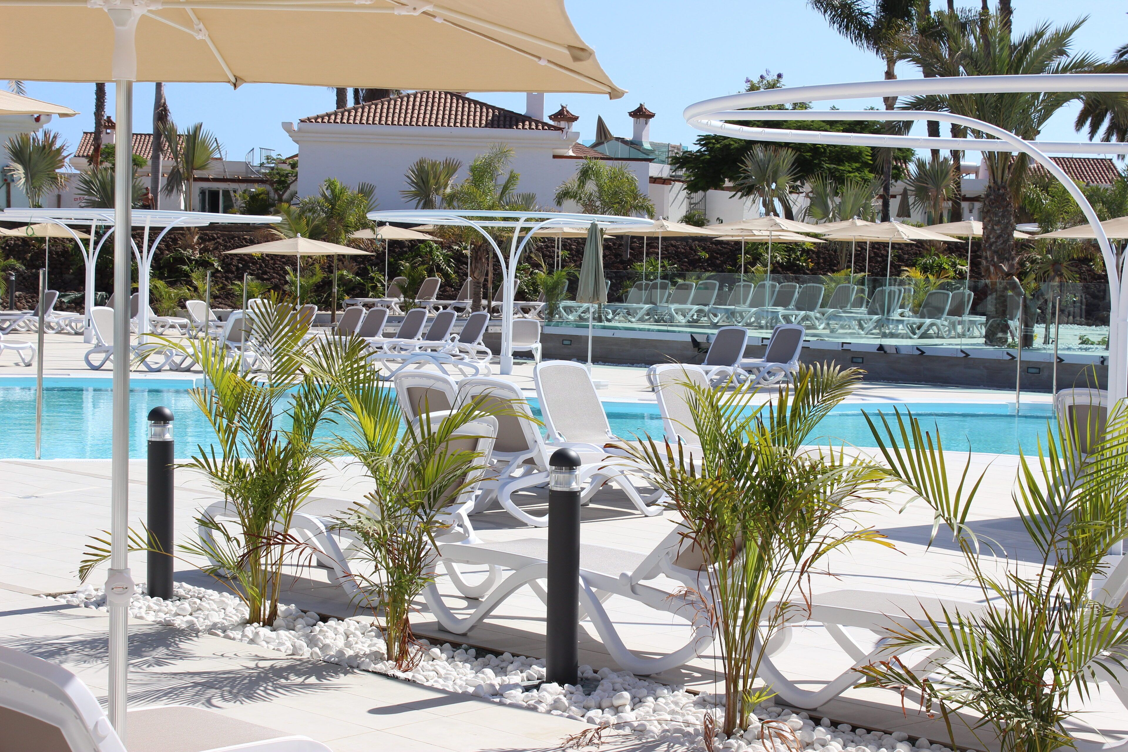 Un área junto a la piscina con sillones blancos, sombrillas y pequeñas palmeras.