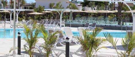 Un área junto a la piscina con sillones blancos, sombrillas y pequeñas palmeras.