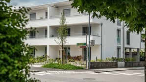 Un edificio moderno con fachada blanca, varios balcones y un pequeño árbol en primer plano.