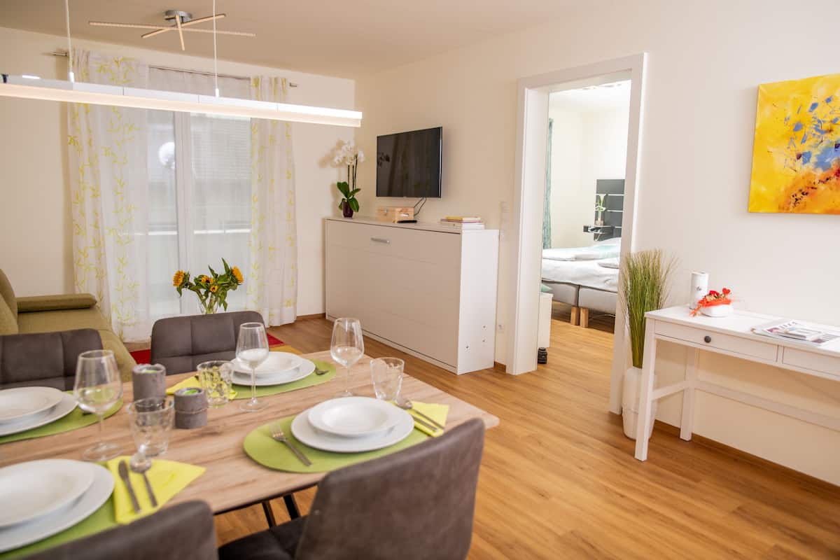 A modern dining area with a wooden table set for four, a flat-screen TV above, and a bright room with a large window.