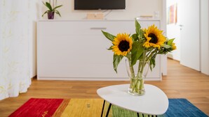 Un salón moderno con un mueble blanco, un televisor de pantalla plana y un jarrón con girasoles sobre una mesa blanca.