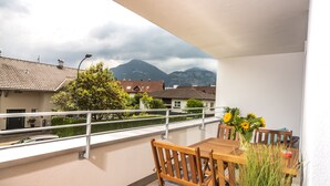 Un balcón con una mesa y sillas de madera, un jarrón con girasoles, y vistas a montañas y casas.