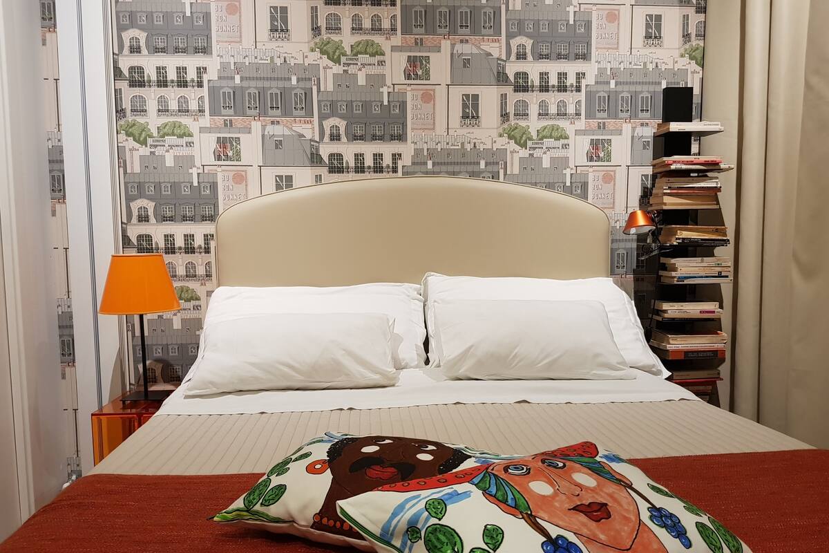 Une chambre d’hôtel avec un lit, des oreillers, une lampe, une bibliothèque et une fenêtre ornée d’un papier peint représentant un paysage urbain.