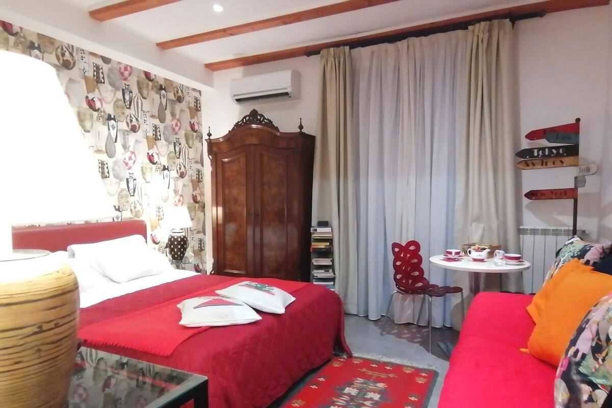 Une chambre d’hôtel avec un lit, un canapé rouge, une petite table avec une théière et un mur décoré de différents objets.