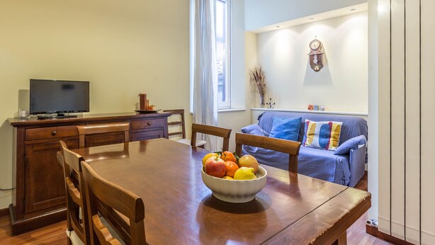 A dining area with a wooden table, chairs, a bowl of fruit, a television on a wooden cabinet, and a blue sofa in the background. - Indipendenza Retreats (Bologna)