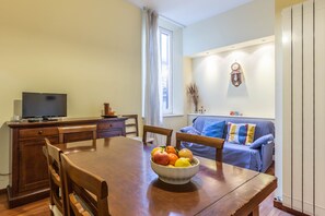 Uma área de refeições com mesa de madeira, cadeiras, uma tigela de frutas, uma televisão sobre um aparador de madeira e um sofá azul ao fundo.