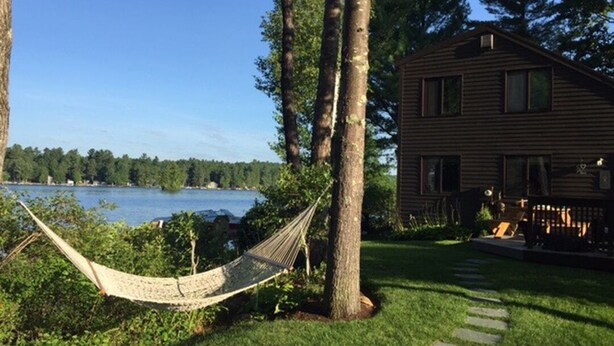 Une cabane en bois avec un hamac, située dans une zone herbeuse près d’un lac.