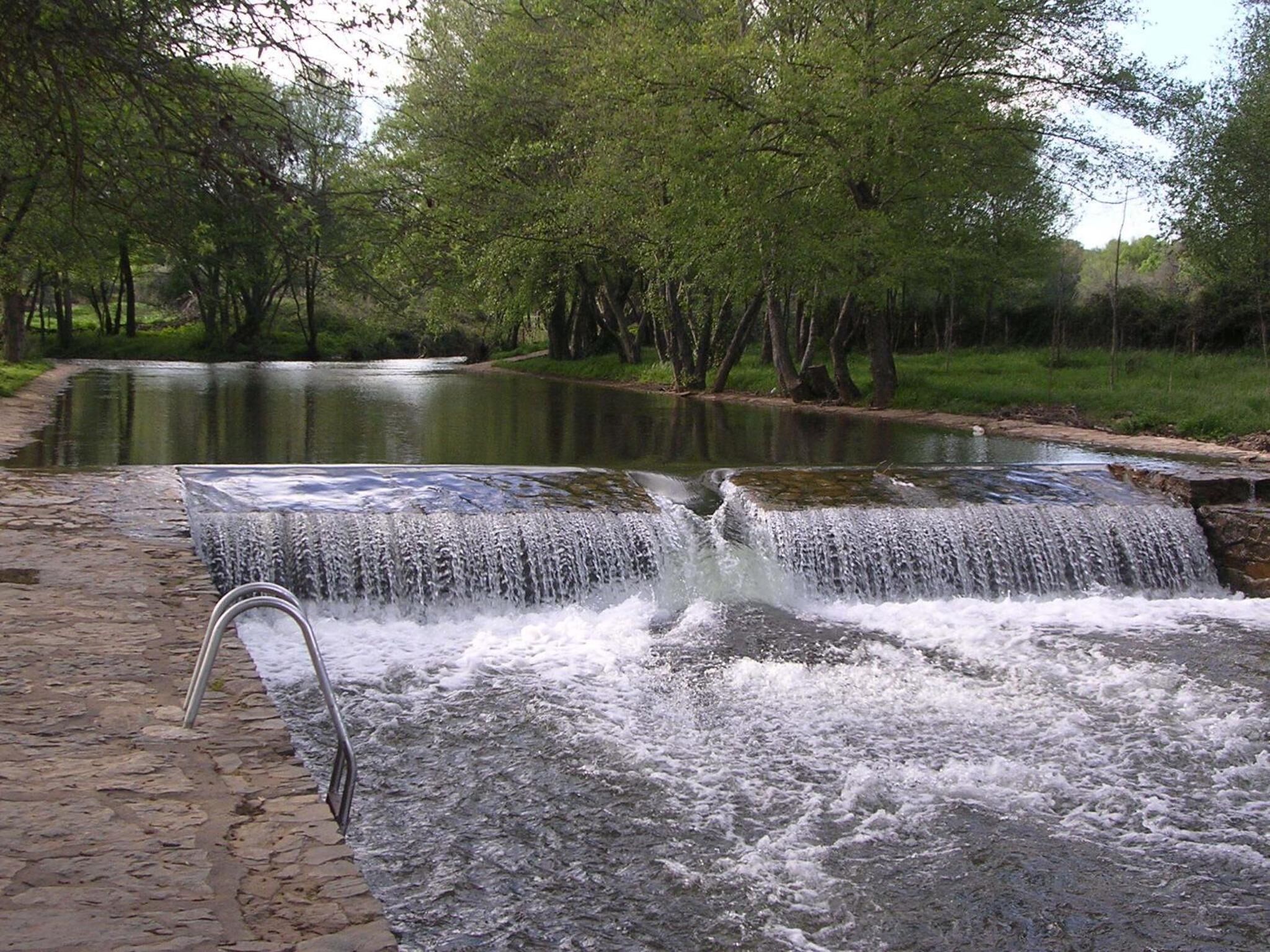 A small waterfall in a park with a paved walkway and trees in the background.