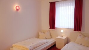 A room with two single beds, a nightstand with a lamp, a window with red curtains, and a wall-mounted light. - Ferienhäuser Seewiesen (Alt Schwerin)