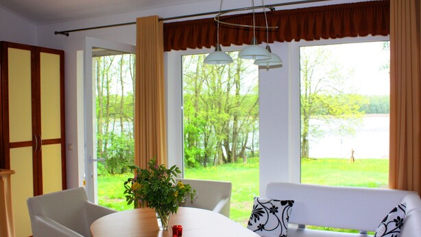 A room with a dining table, chairs, a sofa, and a view of a green landscape through large windows. - Ferienhäuser Seewiesen (Alt Schwerin)