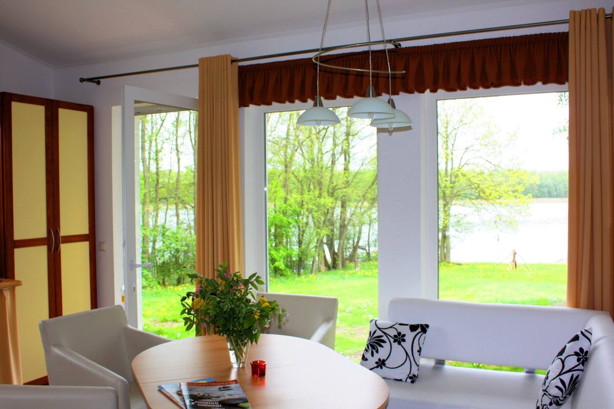 A room with a dining table, chairs, a sofa, and a view of a green landscape through large windows.