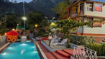 A pool area with a red-roofed structure, a building with a balcony, and a mountainous backdrop.