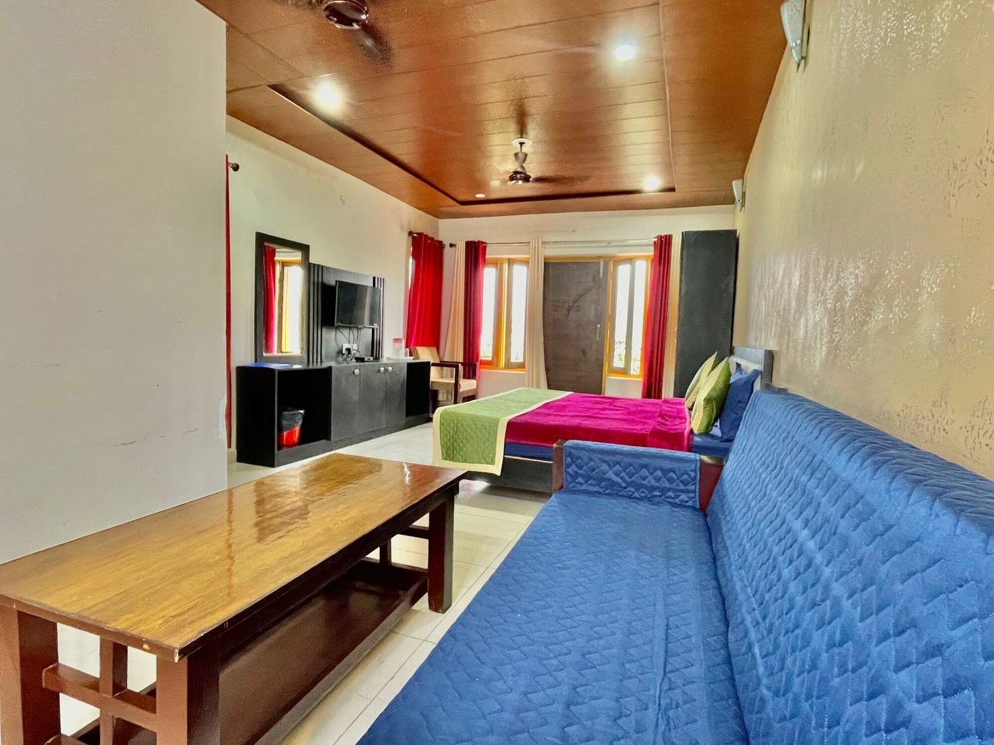 A room with a wooden ceiling, a TV mounted on a black cabinet, a blue sofa, a wooden table, and two beds with colorful bedding.