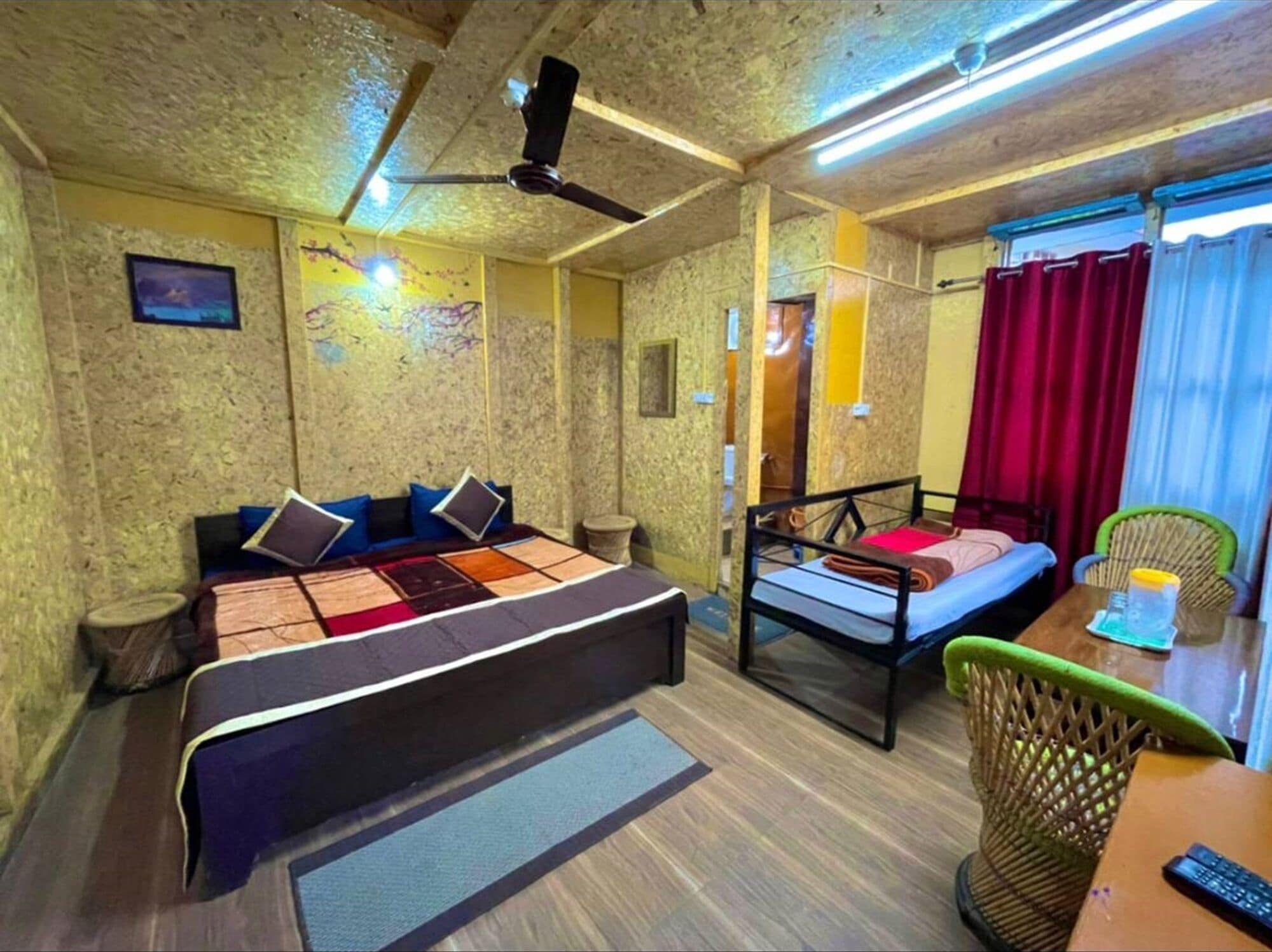 A room with a large bed, a smaller bed, a desk with a chair, a ceiling fan, and a window with curtains.